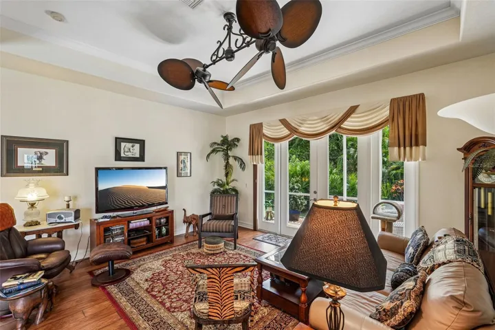 Inviting living area featuring warm wood floors, tray ceilings with crown molding, a dual-head tropical fan, and french doors offering seamless access to lush outdoor views