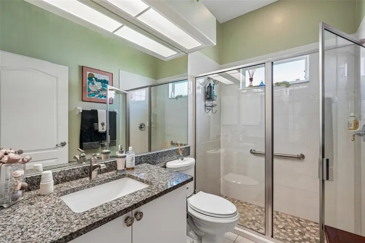Guest bathroom featuring a granite vanity, large mirror with overhead drop-down lighting and a glass-enclosed walk-in shower with river rock tile flooring