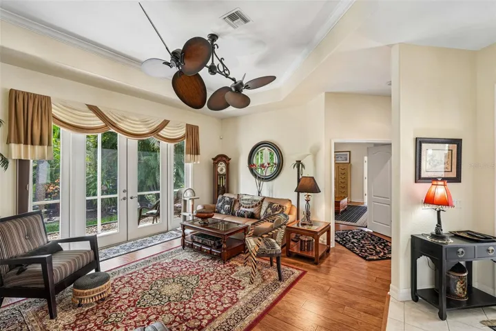 Inviting living area featuring warm wood floors, tray ceilings with crown molding, a dual-head tropical fan, and french doors offering seamless access to lush outdoor views
