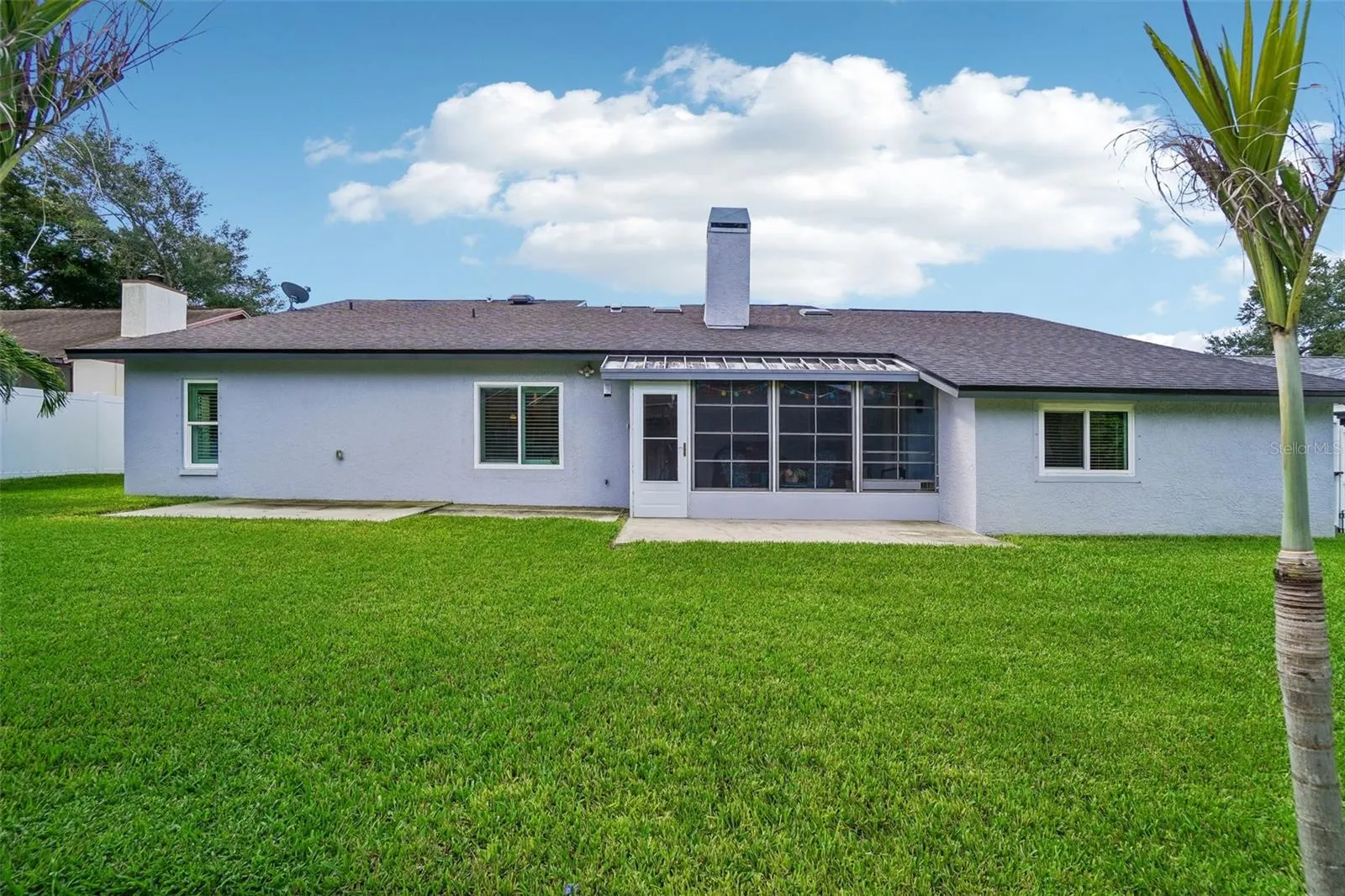 Backyard & Rear Exterior – A straight-on view of the rear elevation showcases the expansive green lawn, enclosed rear patio, and concrete patio areas with mature palms adding a tropical touch.