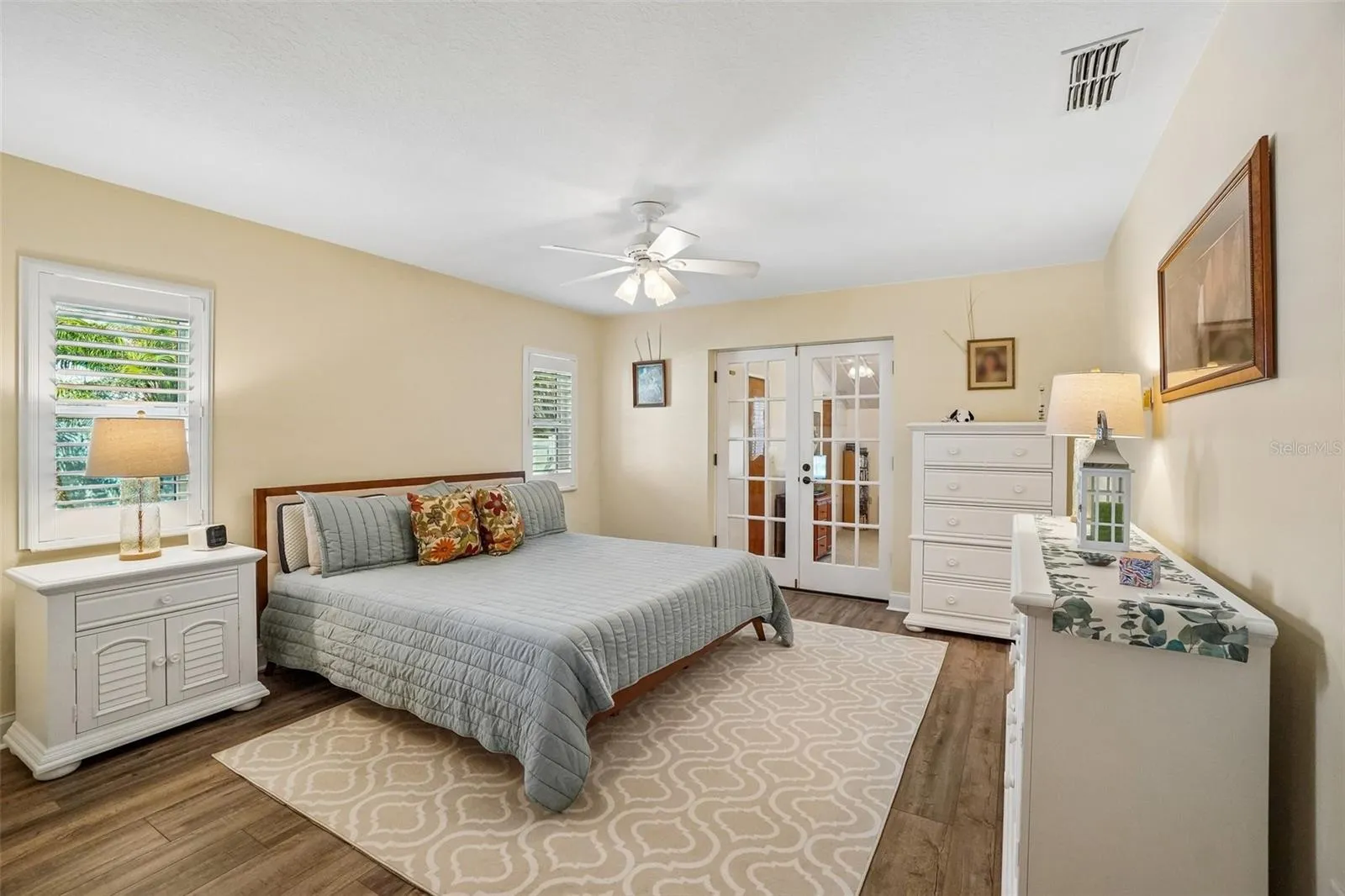 Primary Bedroom – The spacious primary bedroom features wood-look flooring, plantation shutters, a ceiling fan, and French doors that add character while connecting to the adjoining space.