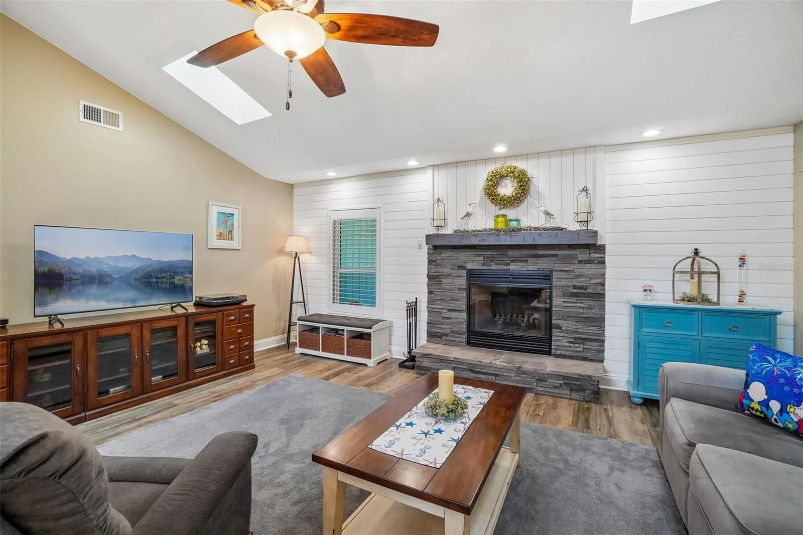 Living Room – A closer view showcases the stacked-stone fireplace and white shiplap accent wall, complemented by wood-look flooring, recessed lighting, and a skylight overhead.