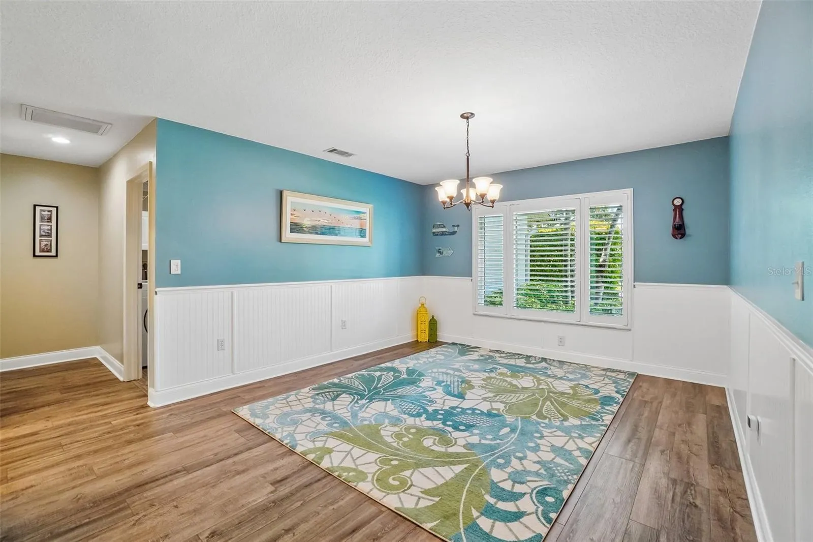 Dining Room – This flexible dining space features wood-look flooring, white beadboard wainscoting, a decorative chandelier, and a large plantation-shuttered window overlooking the greenery outside.