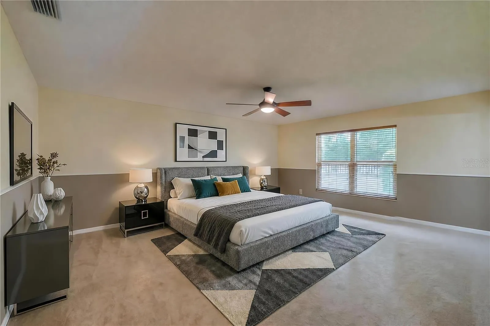 Virtually Staged - Spacious primary bedroom featuring soft carpeting, two-tone painted walls, a central ceiling fan, and a large window providing pleasant outdoor views.
