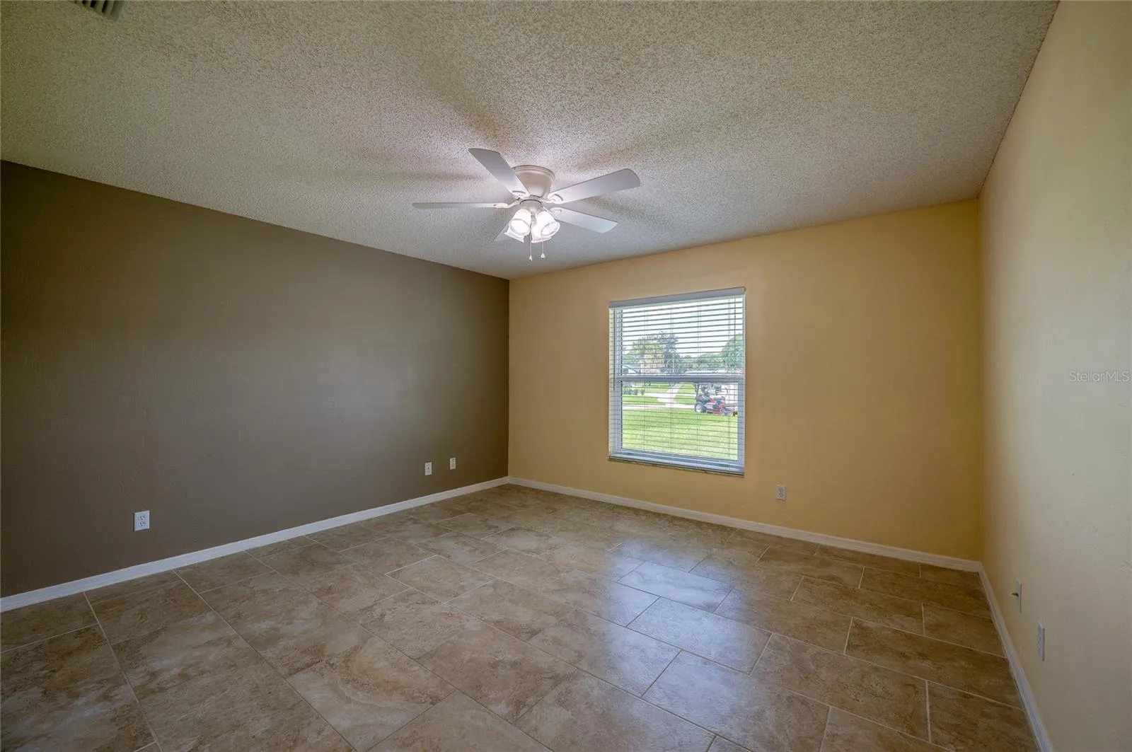 Generous bedroom featuring neutral tiled flooring, dual-tone walls, and a central ceiling fan with integrated lighting. Large window with blinds allows for ample natural light.