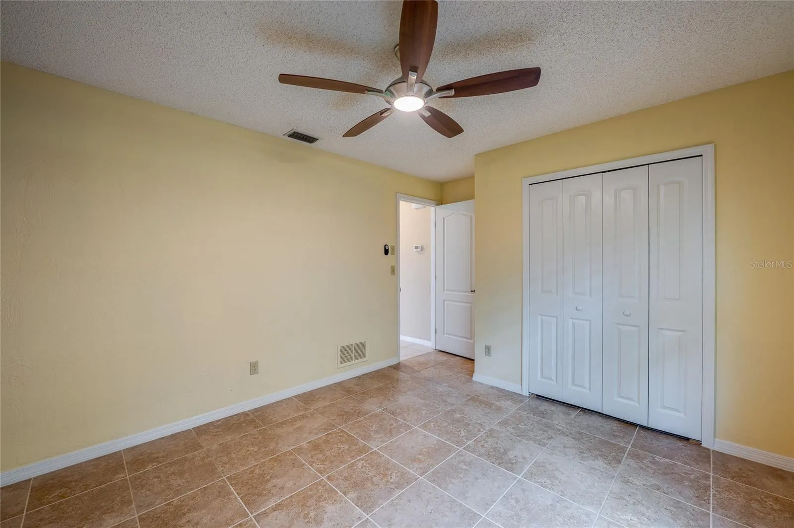 Light-filled bedroom highlighting tiled flooring, smooth neutral walls, and double closet doors for storage. Ceiling fan fixture ensures comfortable airflow.