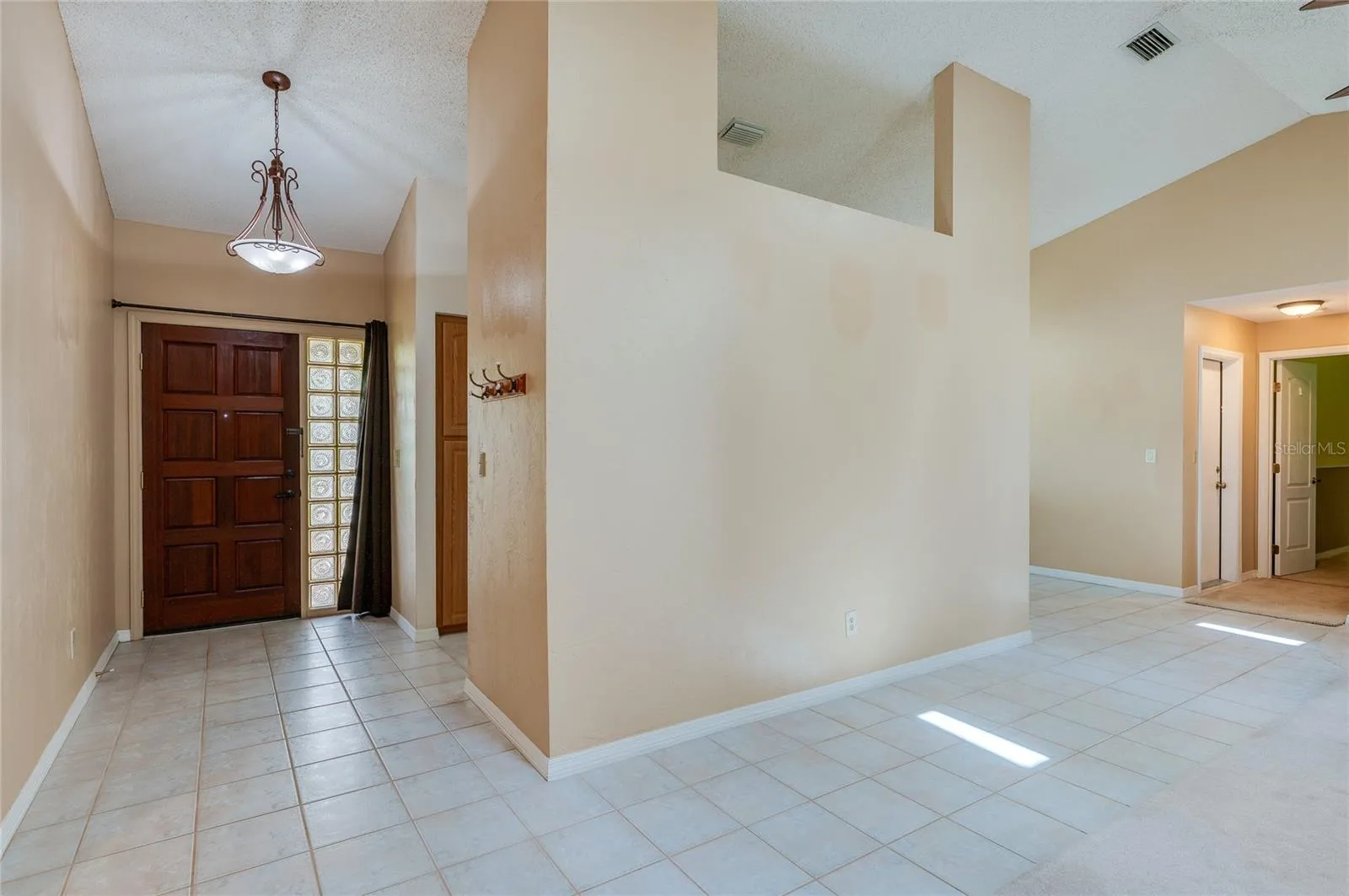 Spacious entry hallway featuring tile flooring, warm neutral walls, and a large decorative wood door with glass block side panels. Vaulted ceilings and open wall cutouts create a light-filled ambiance.