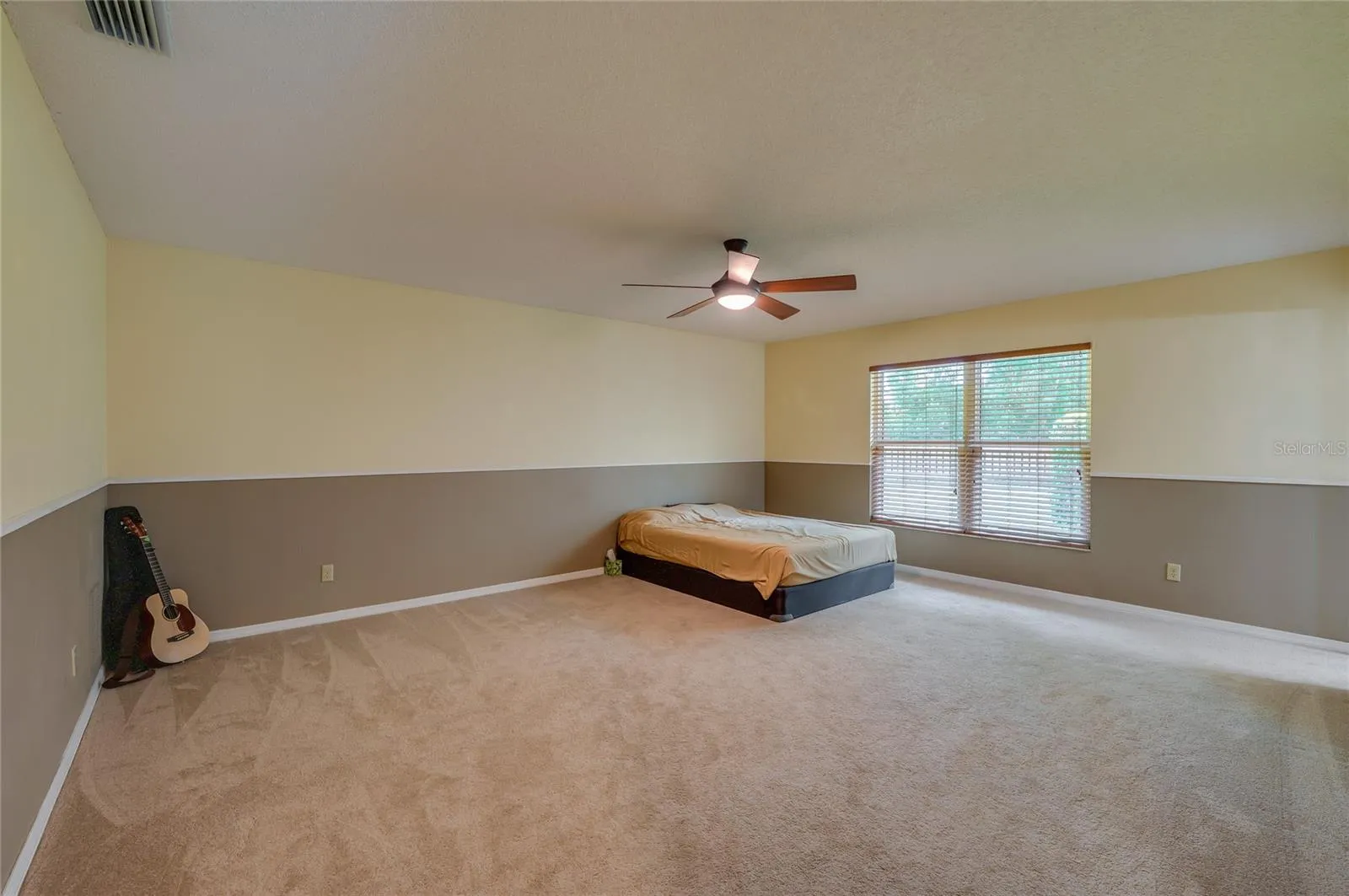 Spacious primary bedroom featuring soft carpeting, two-tone painted walls, a central ceiling fan, and a large window providing pleasant outdoor views.