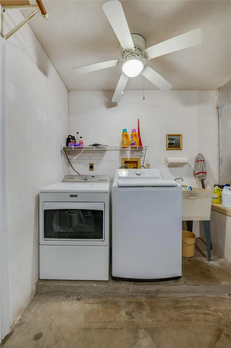 Dedicated laundry area equipped with a side-by-side washer and dryer, utility sink, overhead wire storage rack, and a ceiling fan.