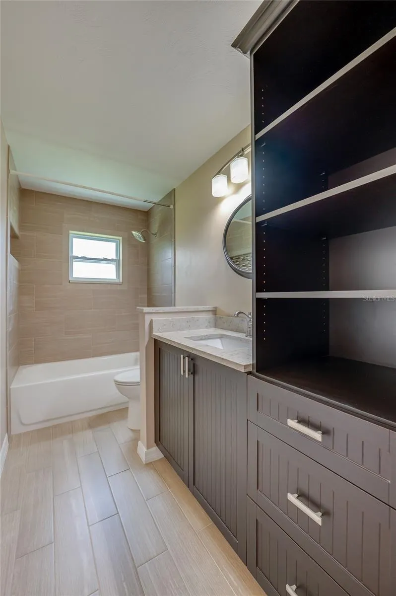 Full bathroom with modern tile flooring, custom dark cabinetry, and open storage. A tiled tub-and-shower combo and elegant wall lighting complete the space.