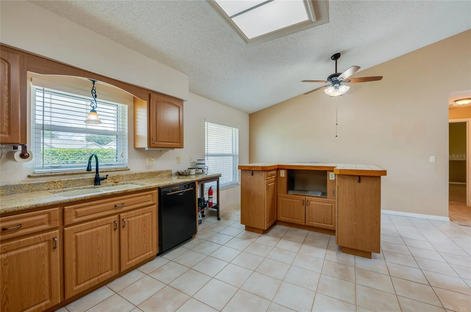 Bright kitchen featuring warm wood cabinetry, neutral countertops, a ceiling fan, and durable tile flooring throughout.