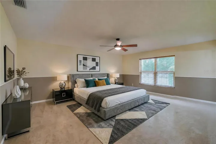Virtually Staged - Spacious primary bedroom featuring soft carpeting, two-tone painted walls, a central ceiling fan, and a large window providing pleasant outdoor views.