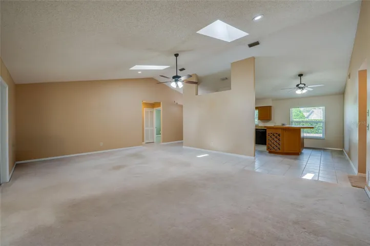 Open-concept living space highlighting high vaulted ceilings, skylights, plush carpeting, and seamless flow into the adjacent tiled kitchen area.