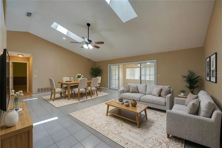 Virtually Staged - Expansive living room area featuring vaulted ceilings with skylights, carpeted flooring, and sliding glass doors that open to a sun room.