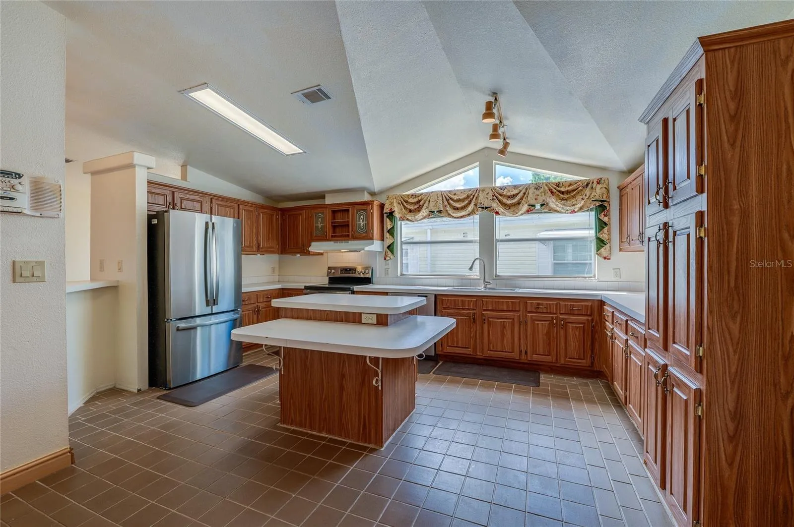 Spacious kitchen features abundant cabinetry, extensive counter space, a center island, stainless steel refrigerator, and expansive windows beneath a vaulted ceiling.