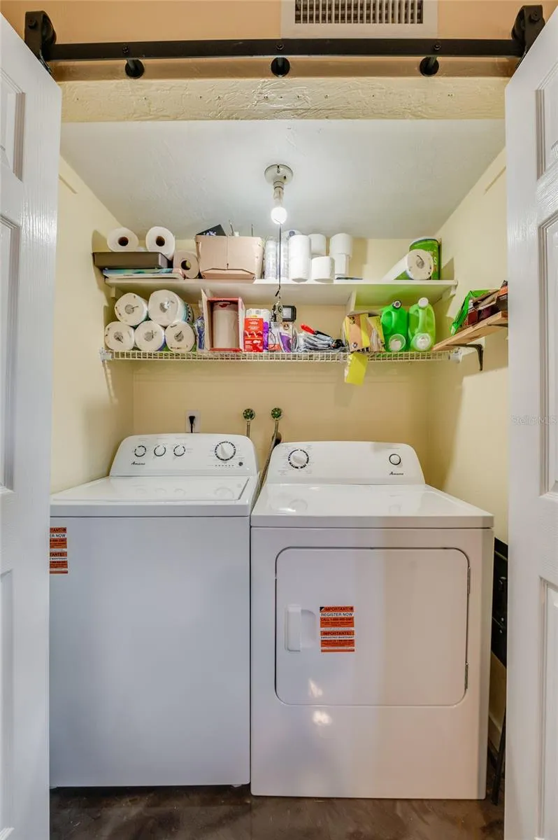 Convenient laundry nook complete with side-by-side washer and dryer units and overhead wire shelving for extra storage.