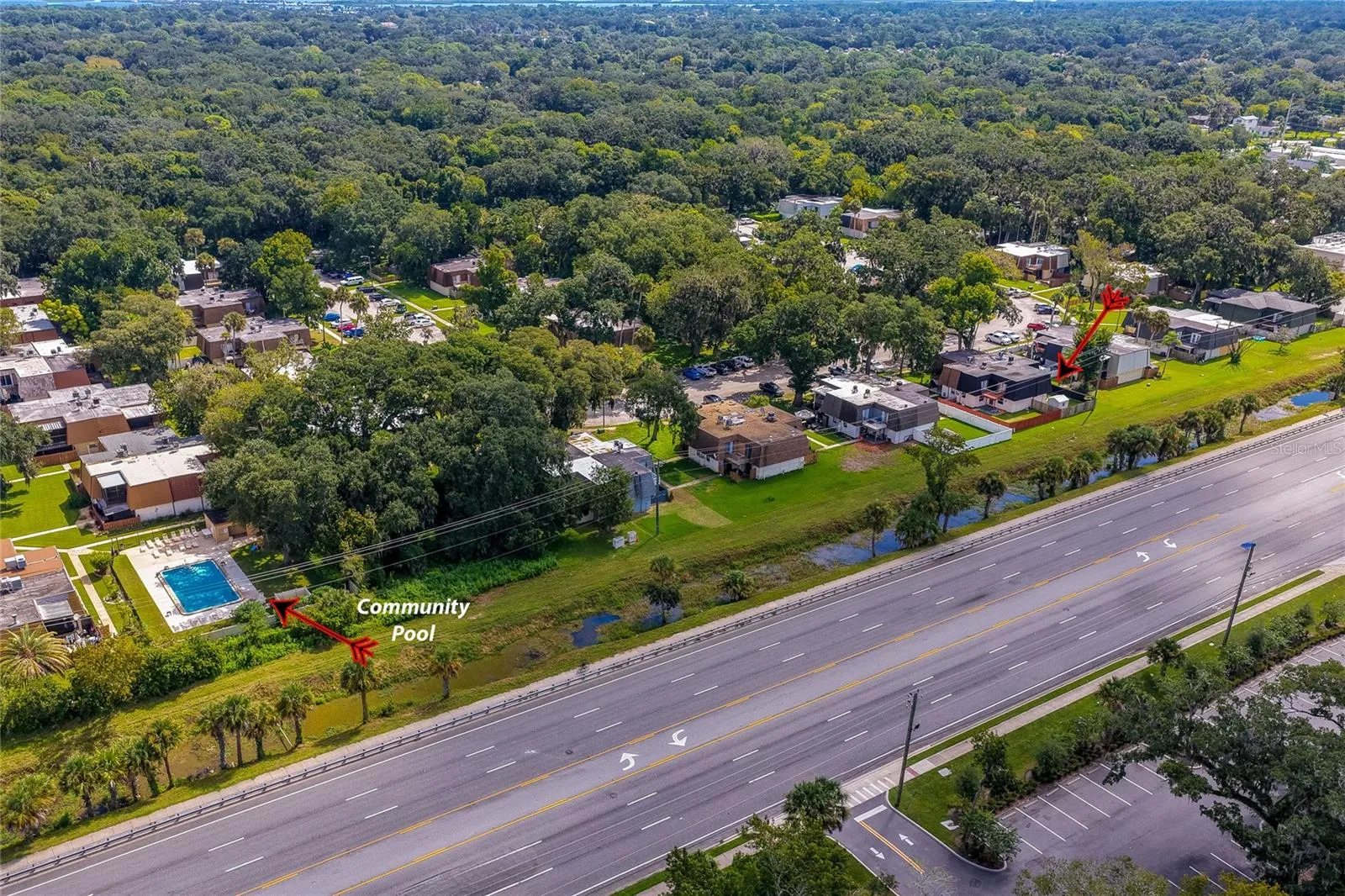 Wide aerial view showcasing the community's layout, highlighting its green surroundings, proximity to the main roadway, and the nearby community pool area.