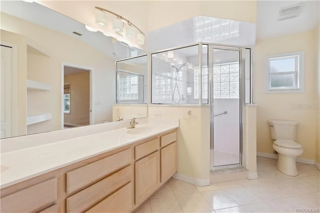 PRIMARY BATHROOM – Wider view showcasing the dual-sink vanity, oversized mirror, glass-enclosed walk-in shower, tile flooring &amp; separate toilet area.