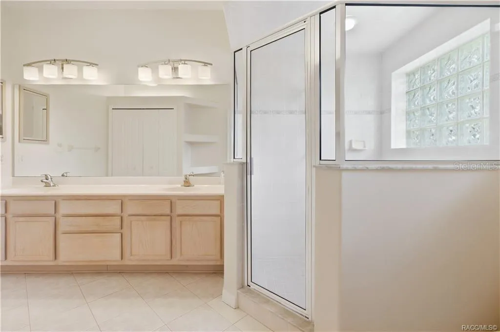 PRIMARY BATHROOM – Primary bath featuring a long dual-sink vanity, expansive wall mirror, tile flooring &amp; large glass-enclosed walk-in shower accented by a glass-block window.