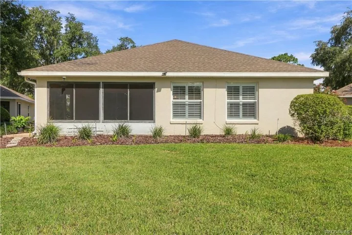 REAR EXTERIOR – Full rear elevation showcasing the enclosed sunroom, additional rear windows with plantation shutters, mature landscaping &amp; spacious green lawn.