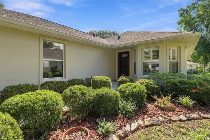 COVERED ENTRY – A closer look at the sheltered front entrance surrounded by mature landscaping. The grouping of front windows with plantation shutters gives the home a polished appearance from both inside &amp; out.