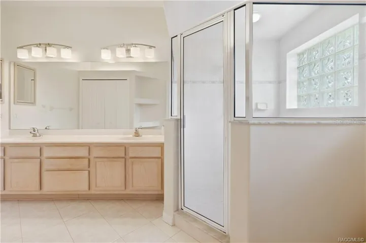 PRIMARY BATHROOM – Primary bath featuring a long dual-sink vanity, expansive wall mirror, tile flooring &amp; large glass-enclosed walk-in shower accented by a glass-block window.