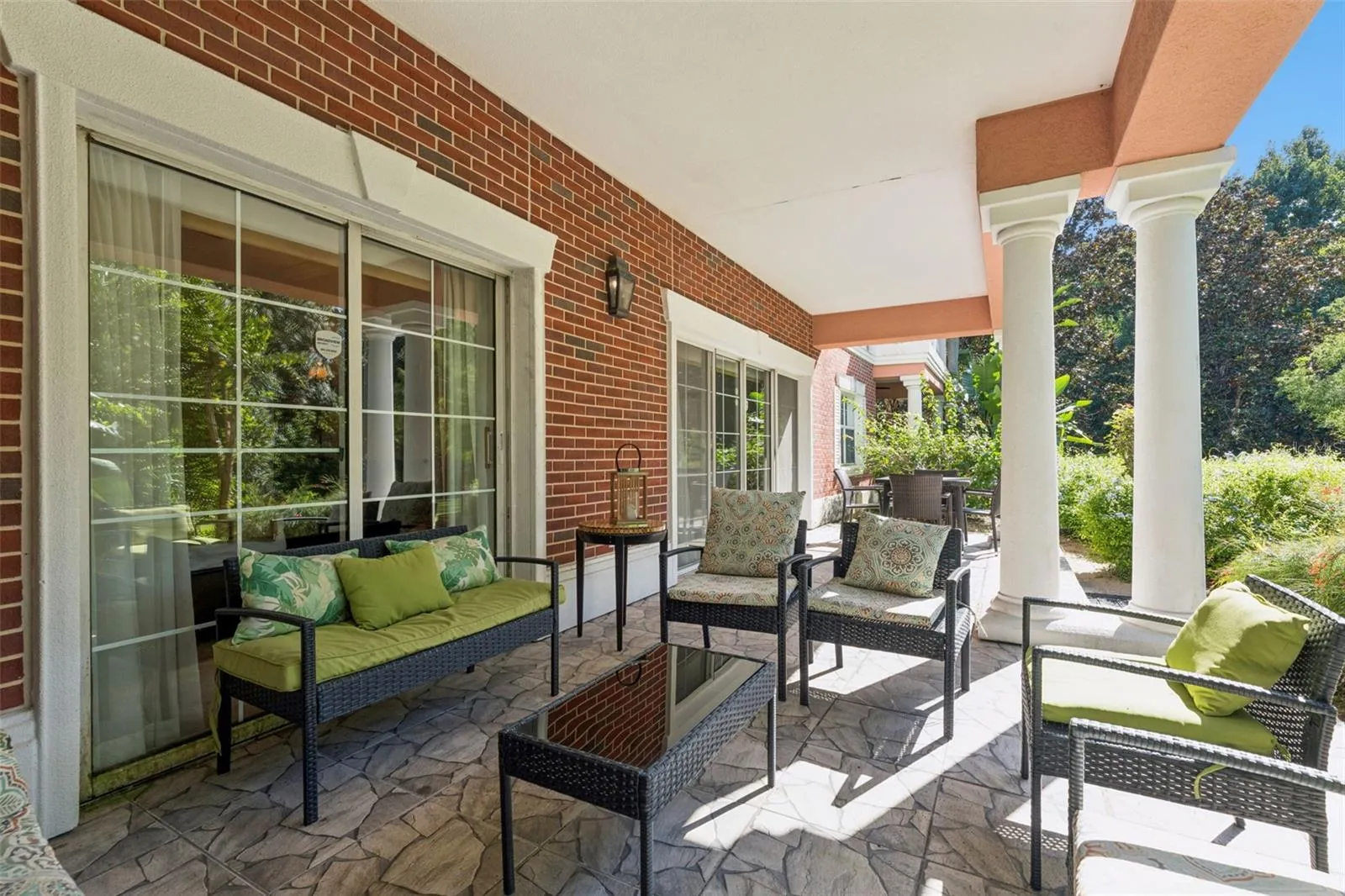 Covered Lanai &amp; Grounds View: Covered porch area featuring white architectural columns, red brick exterior detailing, multiple seating setups, and views of surrounding greenery.