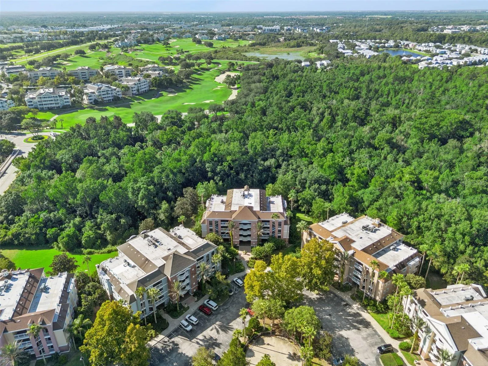 Aerial View of Building &amp; Parking: High-angle aerial view of the condo building nestled directly against a lush conservation backdrop, featuring front parking access and nearby golf course views.