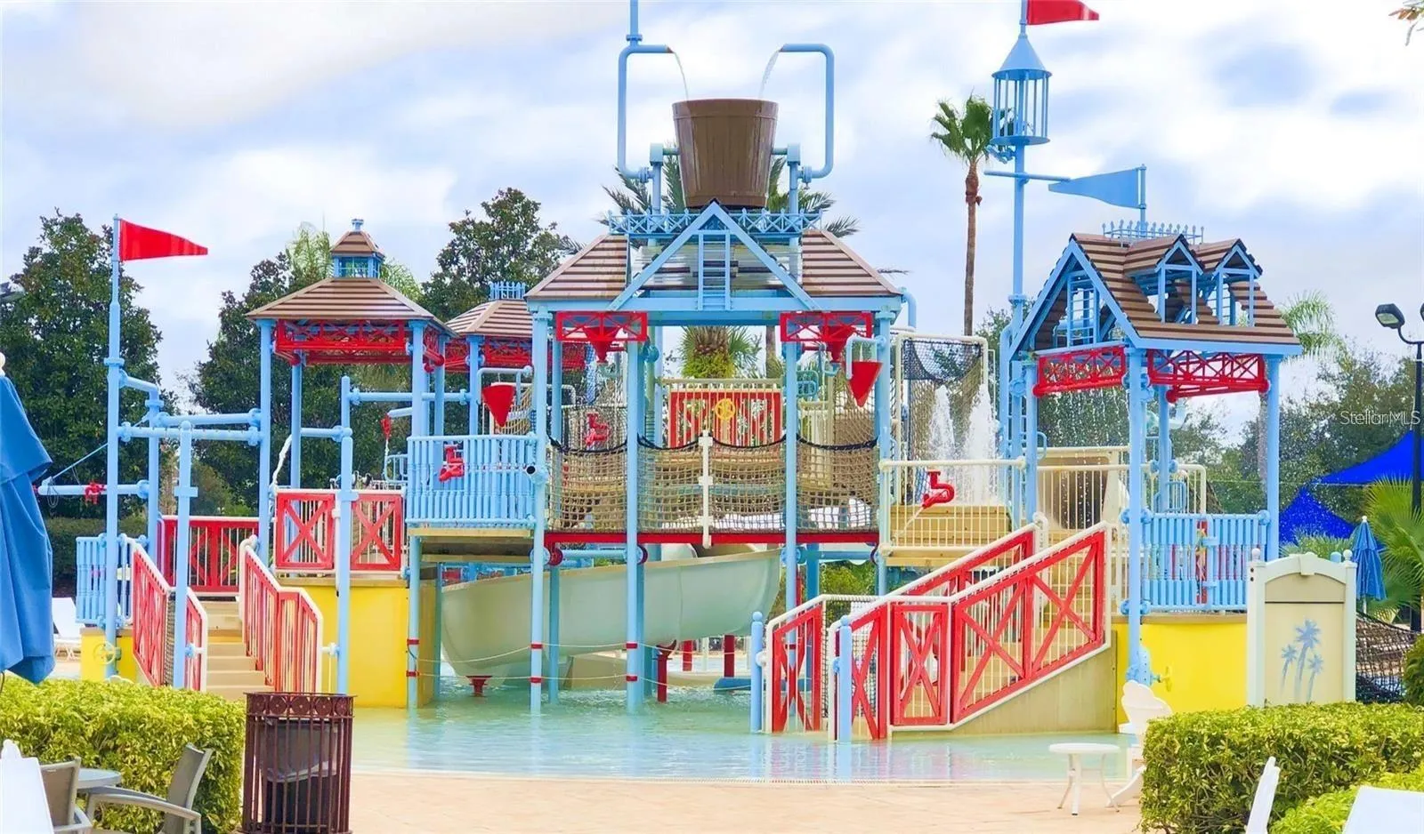 Splash Park Play Structure: Ground-level view of the colorful splash park structure featuring a giant tipping water bucket, slides, and shallow play pool.