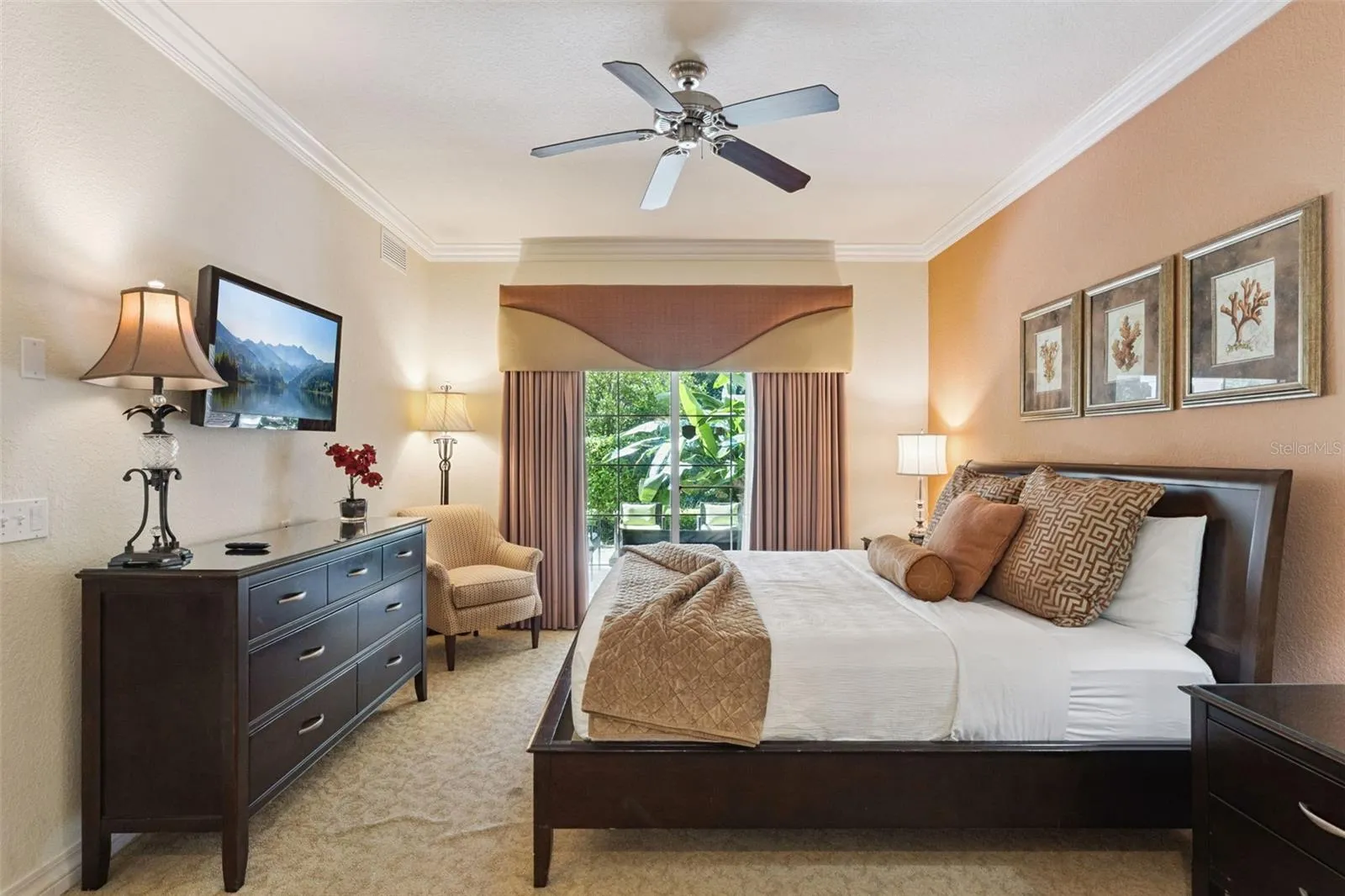 Primary Bedroom: Warmly appointed primary suite featuring crown molding, a ceiling fan, dark wood dresser with wall-mounted TV, cozy seating nook, and sliding glass doors to the outdoor patio.
