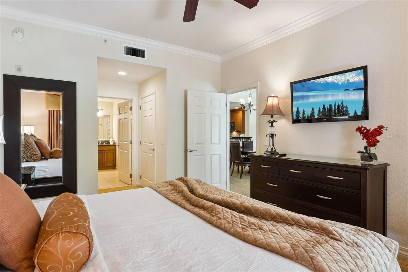 Primary Suite: Spacious primary bedroom featuring crown molding, a ceiling fan, wall-mounted TV, and an ensuite bath entry.