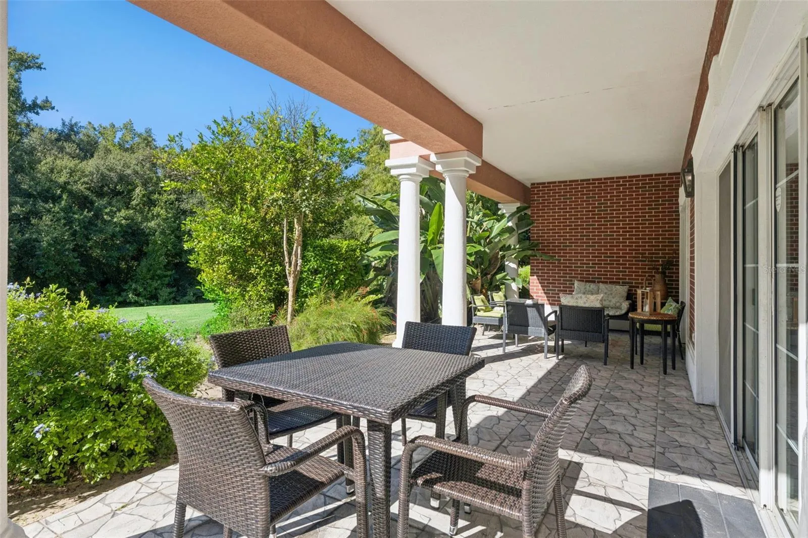 Outdoor Dining Area: Spacious covered patio section featuring a four-person outdoor wicker dining set, white support columns, sliding glass door access, and lush greenery surroundings.