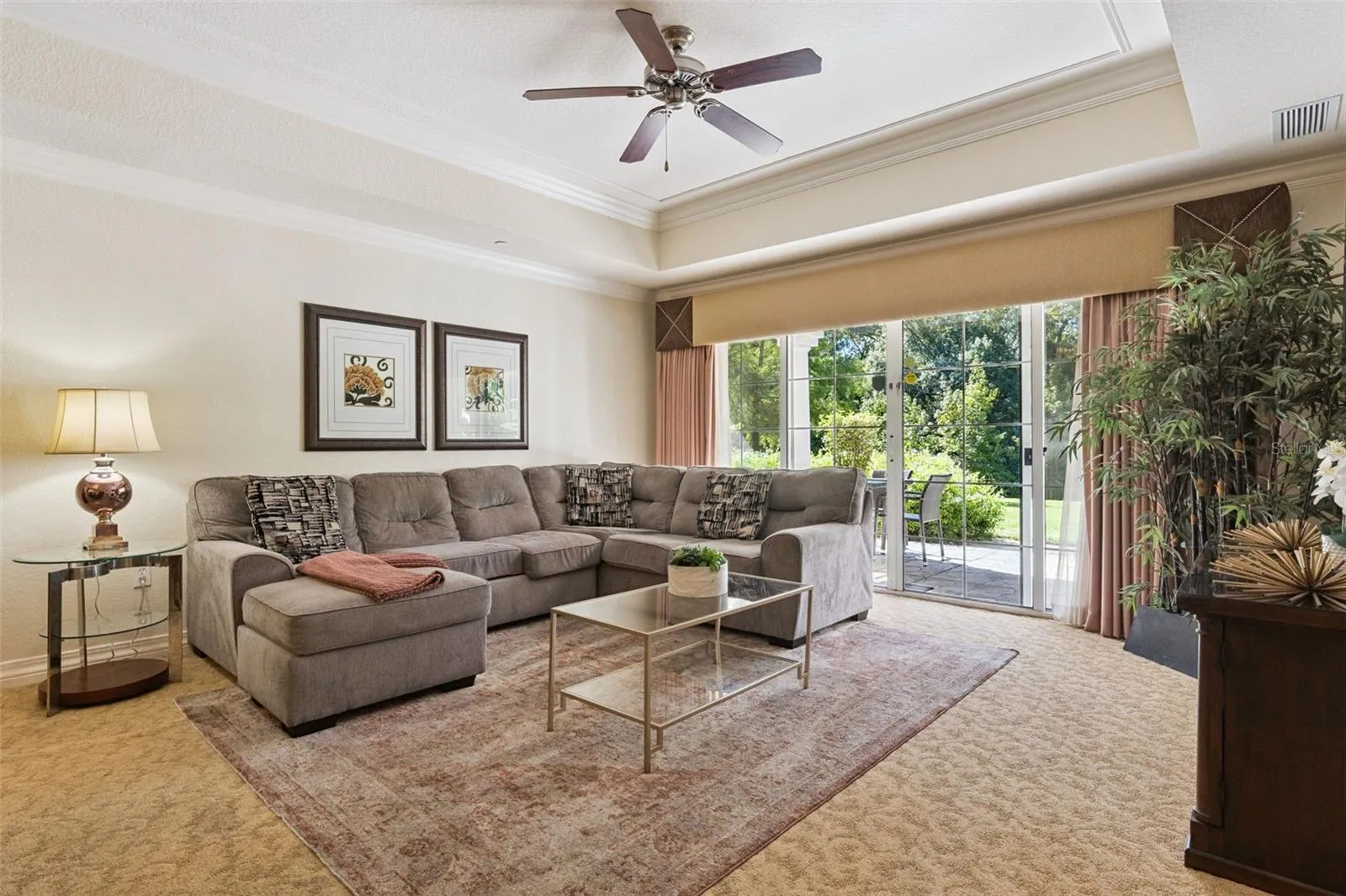 Living Room: Bright living space boasting tray ceilings, plush carpeting, a ceiling fan, and sliding glass doors opening to outdoor greenery views.