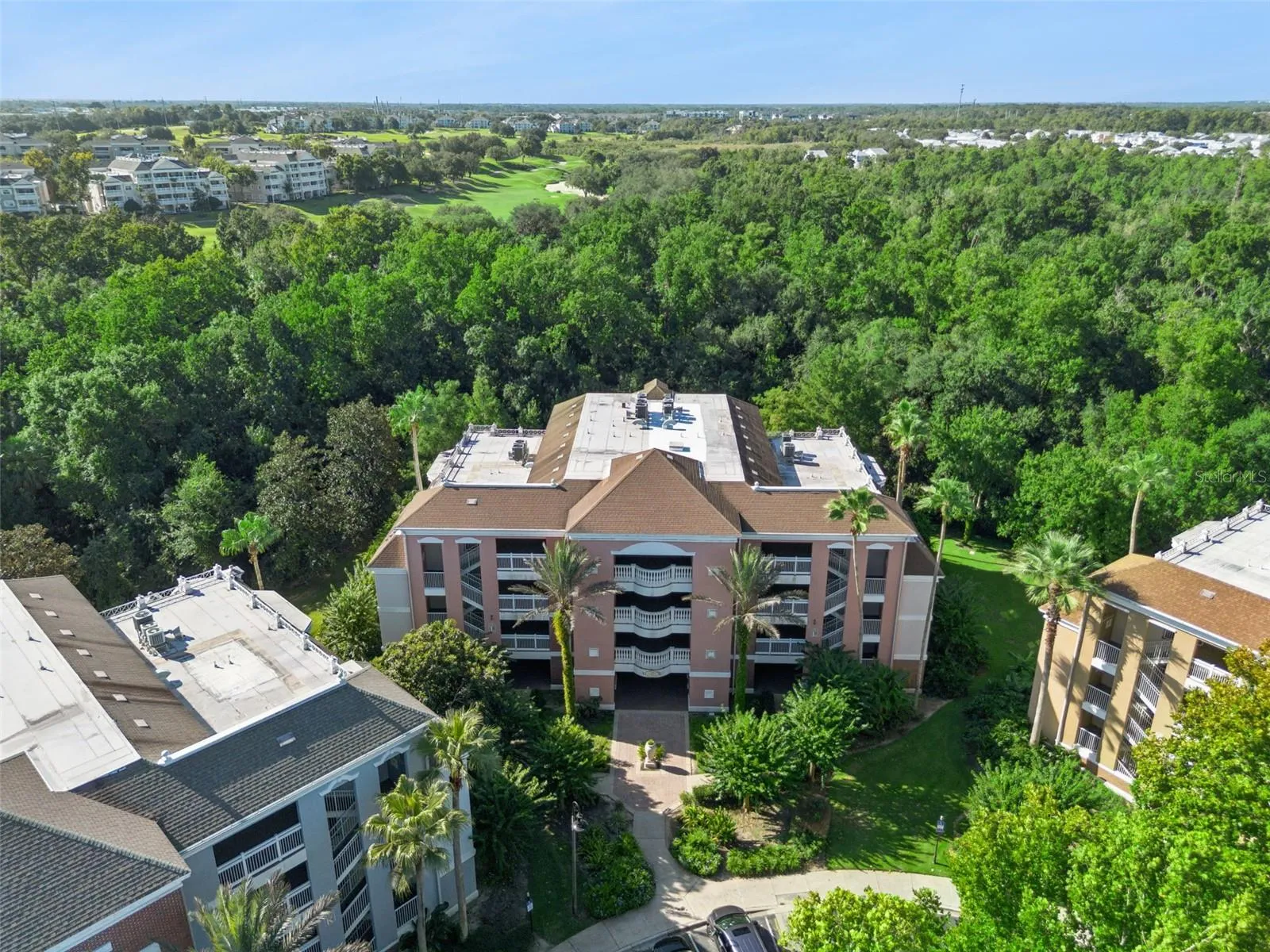 Building Exterior Aerial: Focused aerial shot highlighting the Mediterranean-style building facade, covered central entryway, tropical palm landscaping, and mature tree canopy.