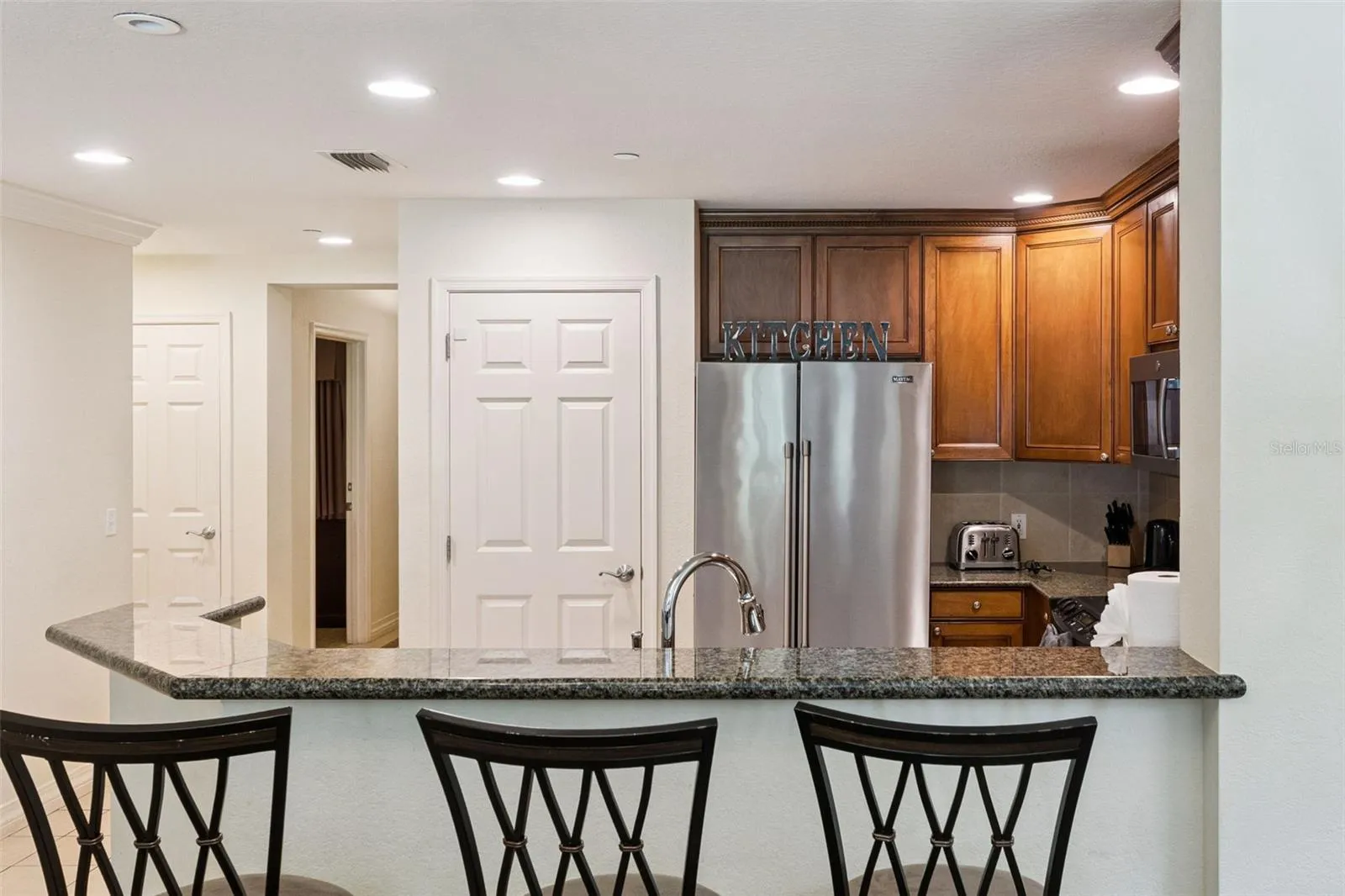 Breakfast Bar &amp; Kitchen: Raised granite breakfast bar ideal for casual seating, featuring a stainless steel refrigerator, recessed lighting, and a pantry door.