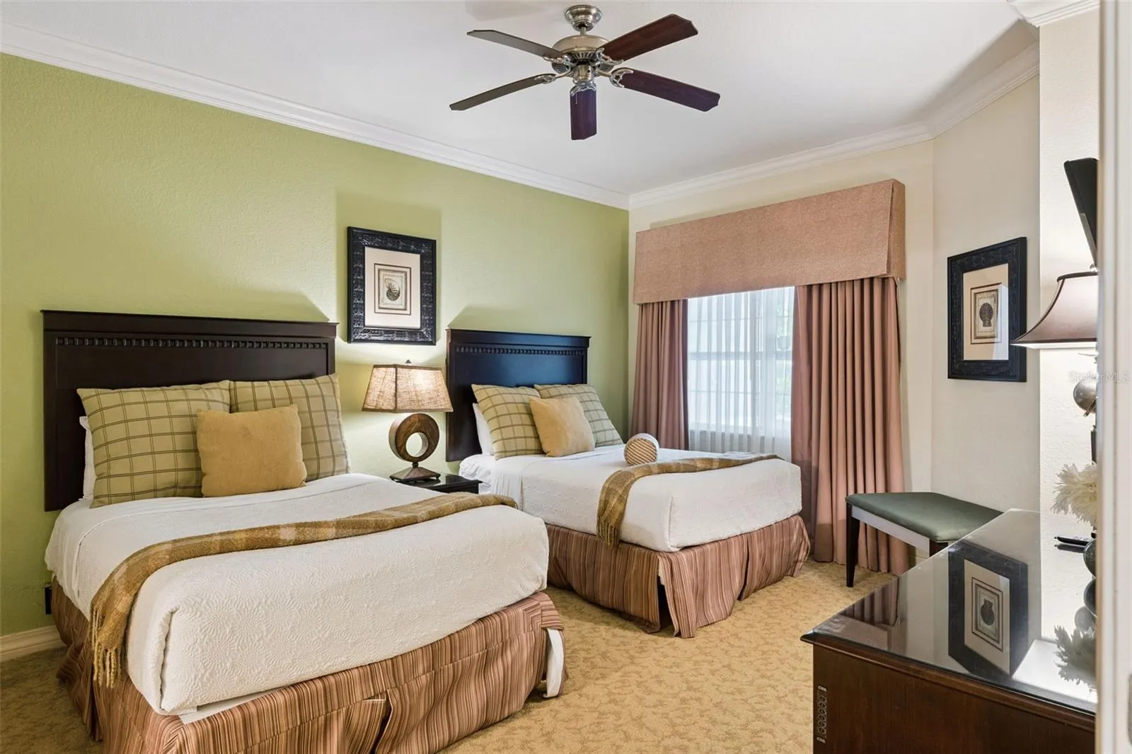 Double Guest Suite: Spacious guest bedroom offering two full beds, a soft green feature wall, central nightstand, ceiling fan, and a large draped window.