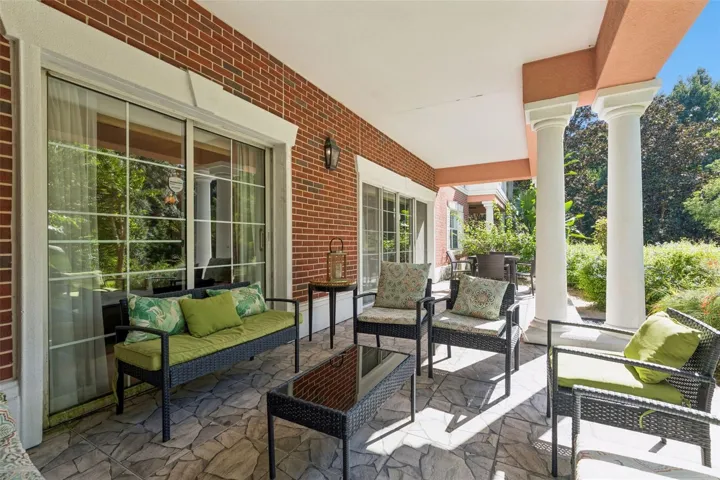 Covered Lanai &amp; Grounds View: Covered porch area featuring white architectural columns, red brick exterior detailing, multiple seating setups, and views of surrounding greenery.