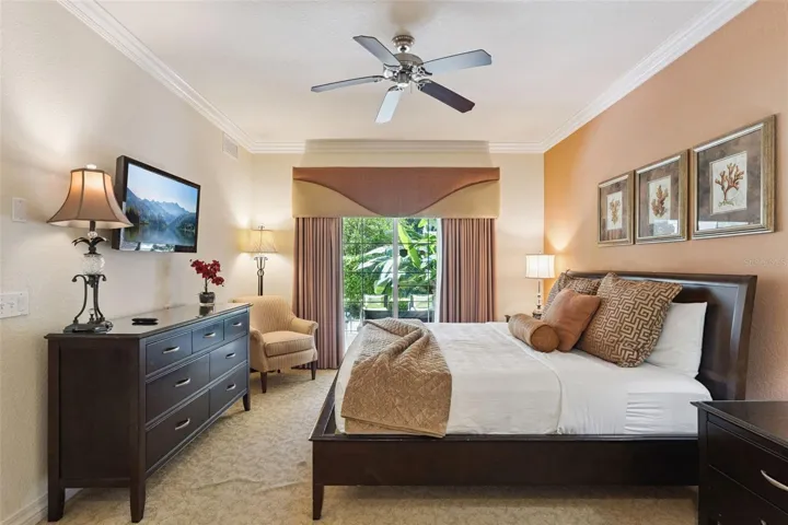 Primary Bedroom: Warmly appointed primary suite featuring crown molding, a ceiling fan, dark wood dresser with wall-mounted TV, cozy seating nook, and sliding glass doors to the outdoor patio.