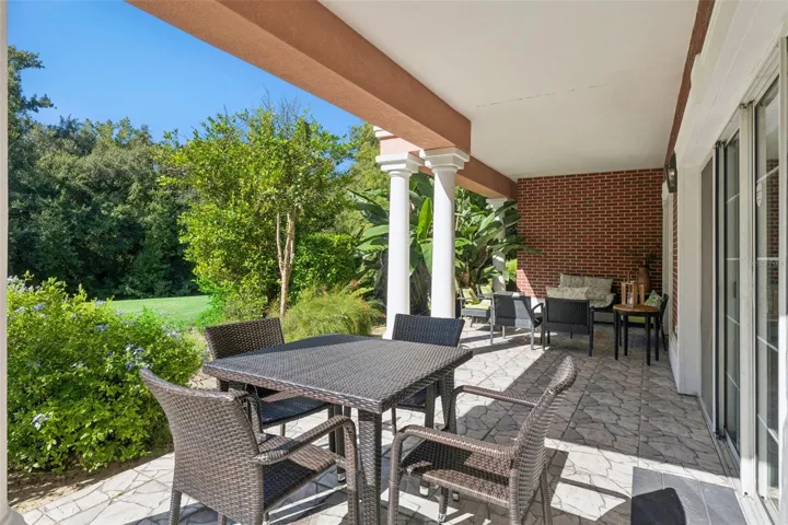Outdoor Dining Area: Spacious covered patio section featuring a four-person outdoor wicker dining set, white support columns, sliding glass door access, and lush greenery surroundings.