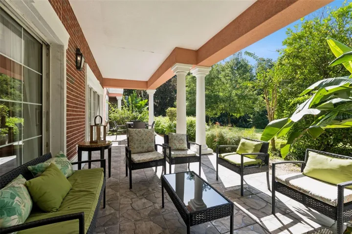 Covered Patio Seating: Covered outdoor patio with stone-patterned tile flooring, classic red brick wall accents, comfortable wicker lounge seating, and views of surrounding tropical flora.