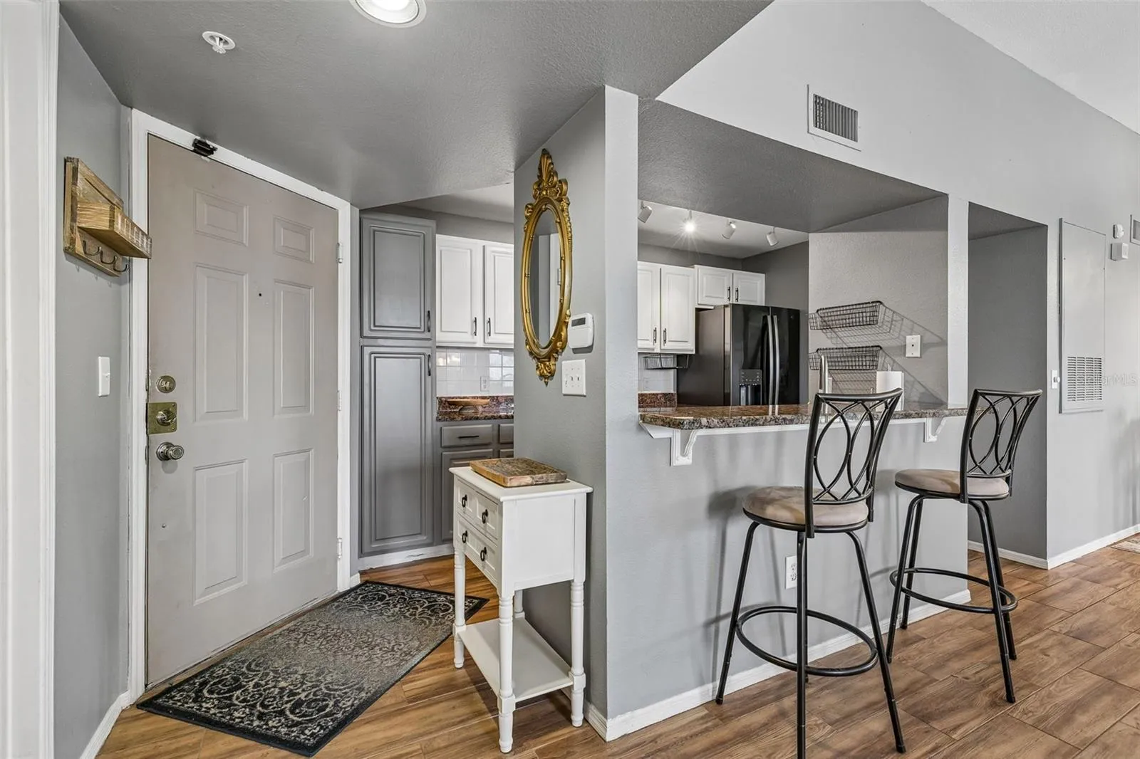 Entry foyer of Unit 314 looking toward the kitchen breakfast bar with two counter stools, white cabinets, and ceramic tile wood-look plank flooring.