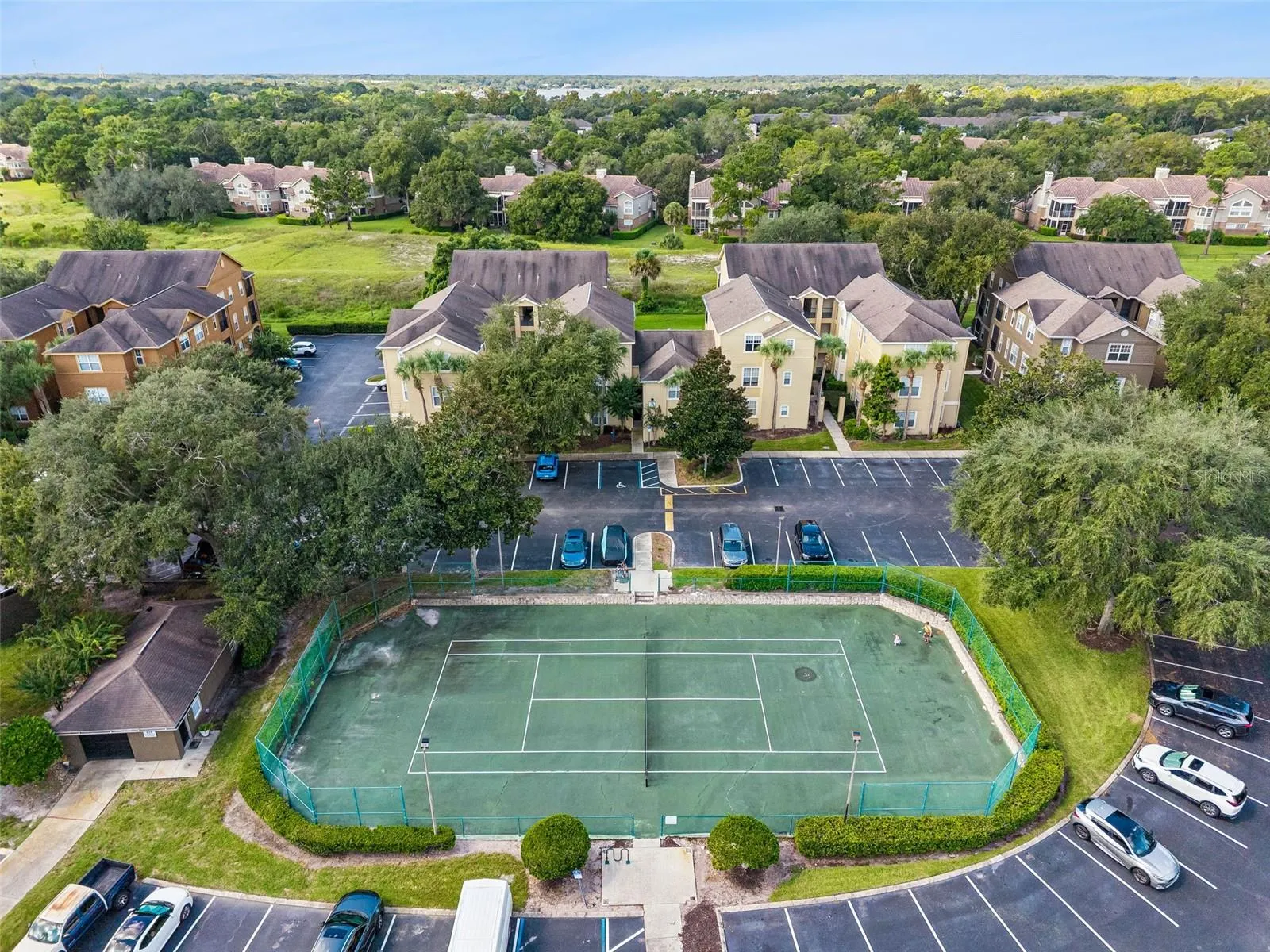 Sabal Point tennis court with green playing surface and fencing, next to a parking area and residential buildings.