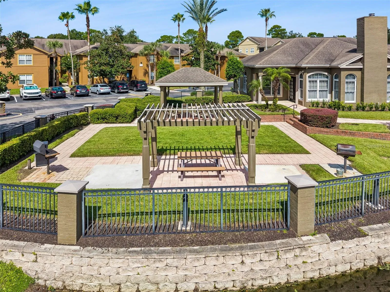 Sabal Point pergola with a picnic table and barbecue grills on a brick paver patio beside the lake wall, with the clubhouse in the background.