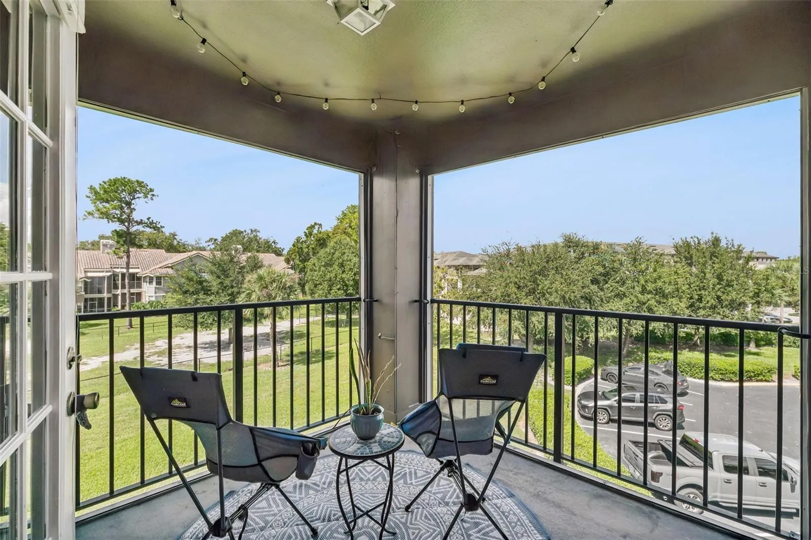 Screened balcony with two chairs, a small table, and string lights, overlooking green space and a parking area below.