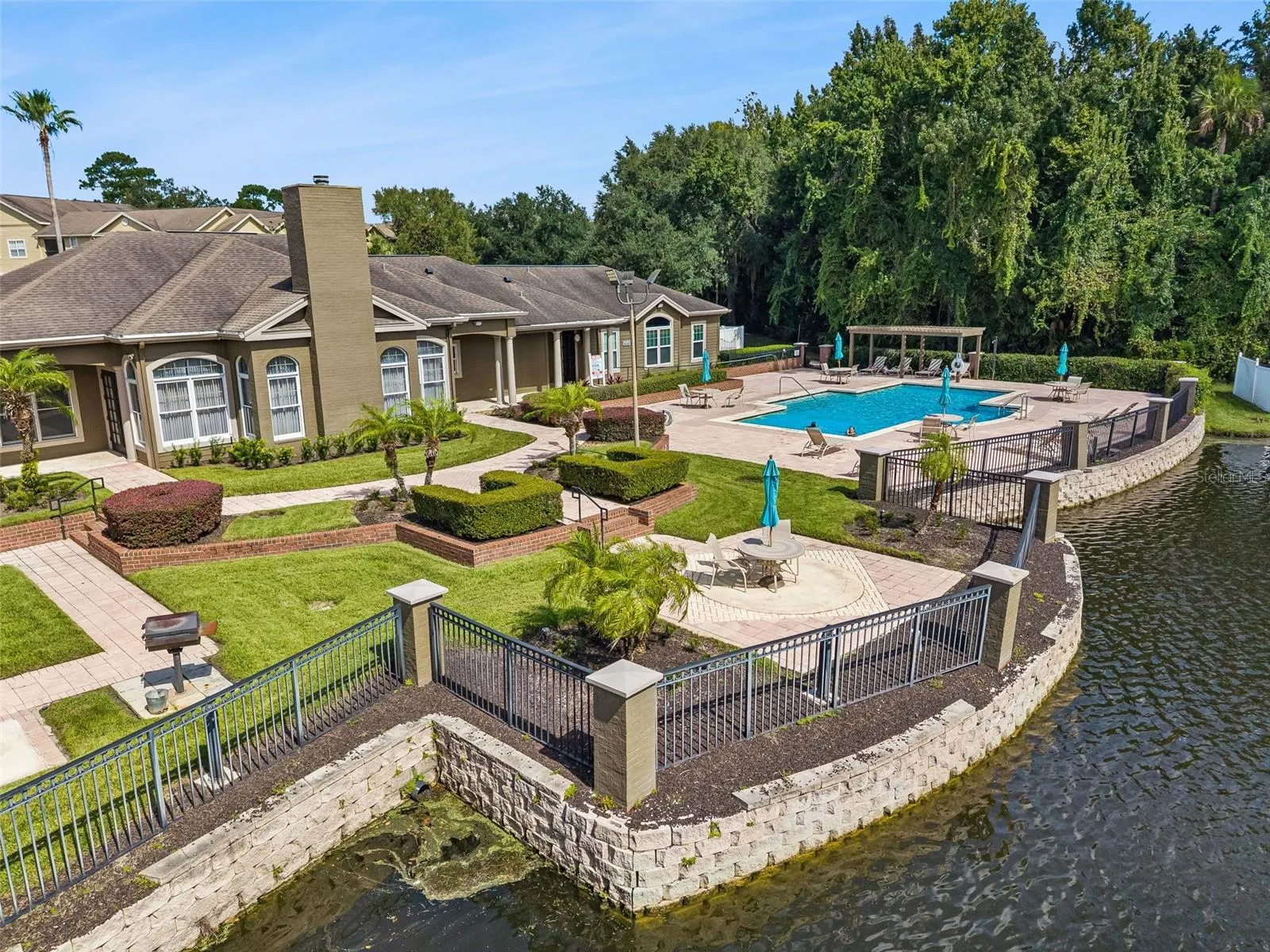 Aerial view of the Sabal Point clubhouse and pool from the lake side, with a stone retaining wall and black metal fencing.
