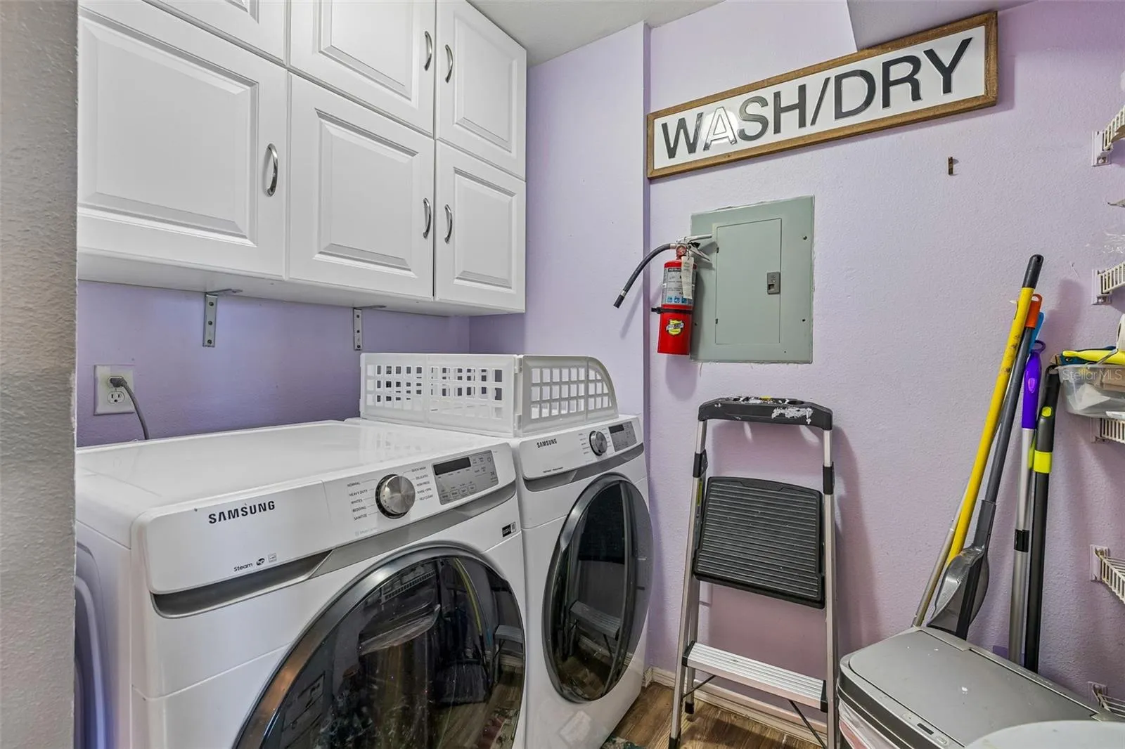 Laundry closet with a Samsung front-load washer and dryer, upper white cabinets, shelving, and the electrical panel.