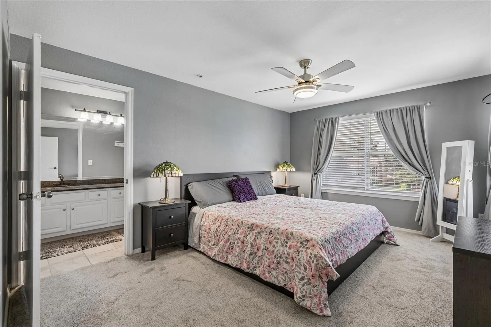 Primary bedroom with a king bed, carpeted floor, ceiling fan, window with curtains, and a doorway to the en suite bathroom.