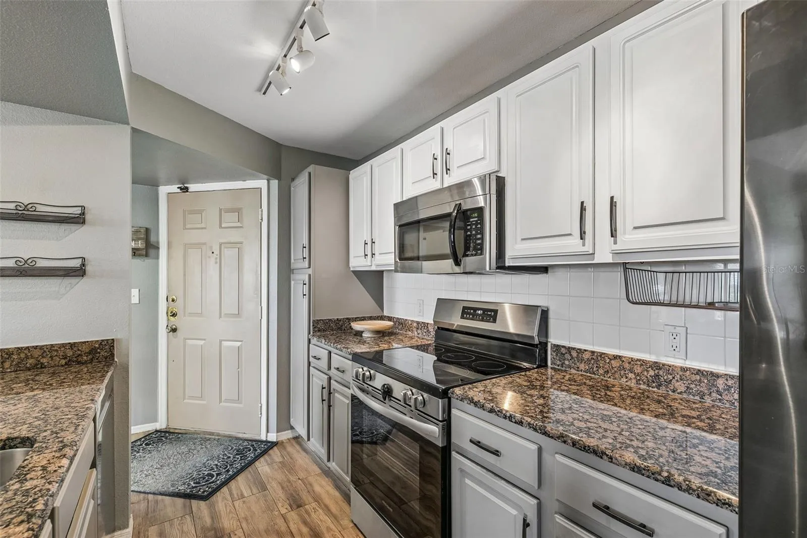 Kitchen with white raised-panel cabinets, granite counters, glass-top electric range, stainless microwave, and track lighting, with the entry door in the background.