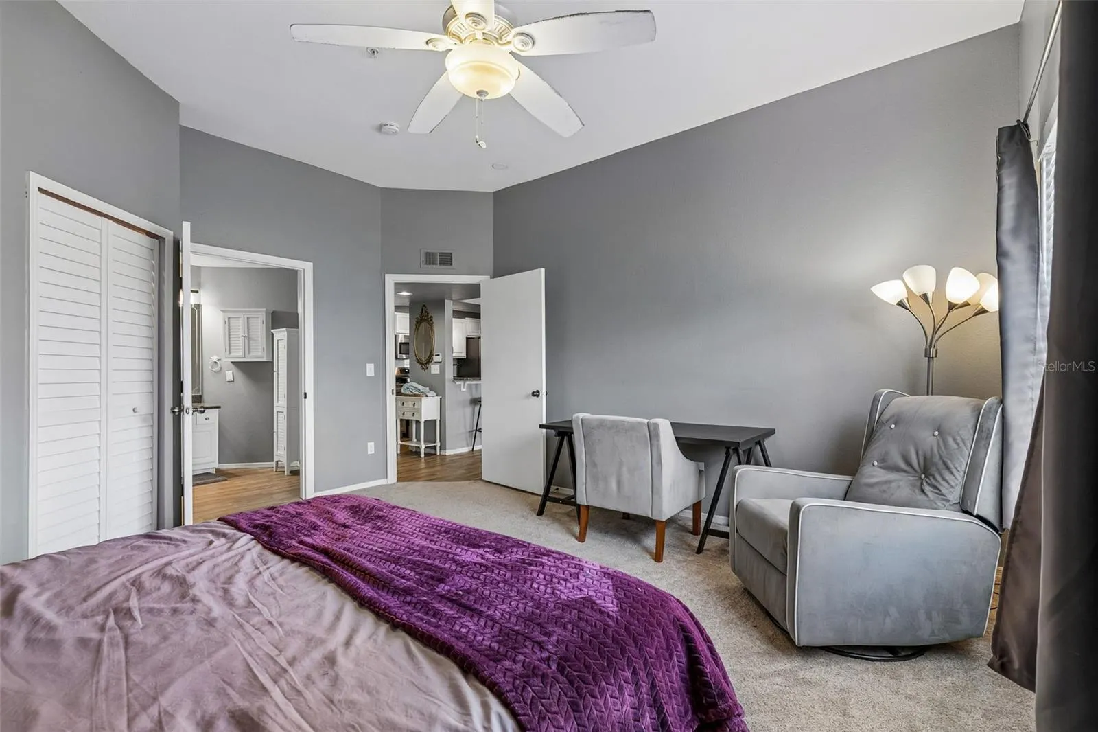 Second bedroom sitting area with a desk, accent chair, ceiling fan, and louvered closet doors.