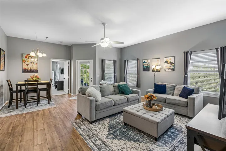 Living room and dining area in an open layout with a chandelier, ceiling fan, ceramic tile wood-look flooring, and windows on two walls.